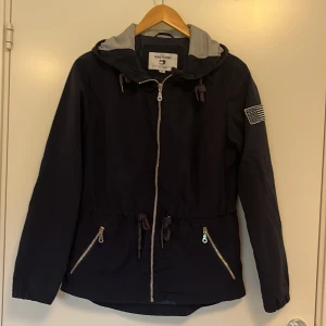 Mörkblå jacka i nylon och polyester  - Jackan är mörkblå med silvriga detaljer såsom dragkedja och flagga på höger arm. Det går att ställa in jackan i midjan och vid luvan. Luvan har blå och vit randigt innertyg. OBS! Jackan är inte fodrad 
