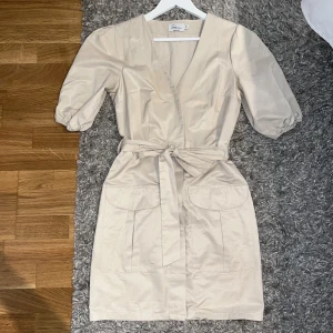 Klänning med knytning  - En riktigt snygg beige klänning med  knytning i midjan och fickor på låren. Den är 88cm från axlar till nerkant. Den kommer från Sara Sieppis kollektion tillsammans med NA-KD 