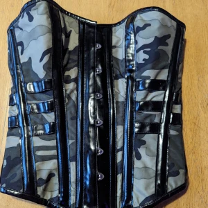 Camo steel boned overbust corset 26" - En korsett med camouflage mönster ifrån Glamorose. Har en en skada då sömmen har släppt på ena sidan av stängning med planchetter, dock på undersidan så detta syns inte när man bär korsetten (kan skicka bilder) Storlek M vilket motsvarar 76-90cm midja