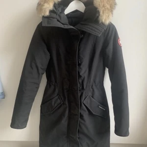 Canada Goose Rossclair Parka - Fin jacka från Canada Goose i modell Rossclair. Medellång jacka med päls på luvan som man kan ta bort och den är justerbar i midjan.  Svart. Canada Goose märke på ena ärmen.  Storlek XXS.  Den är köpt på NK i Stockholm, kvitto finns. Använd 2 säsonger men sparsamt.