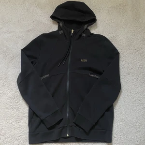 HUGO BOSS - Skön hoodie/huvtröja med avslappnad passform. Guldiga mönster vid kragen och under brösten, samt guldig dragkedja. Fickor vid sidorna