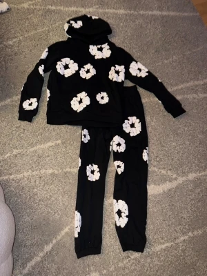Svart mjukisset från denim tears - Mysigt svart mjukisset från denim tears. Setet består av en hoodie och ett par byxor, båda med samma blommönster. Perfekt för hemmamys eller som bekväma vardagskläder. Använd men i fint skick, det står att det är storlek L men passformen är för storlek M.