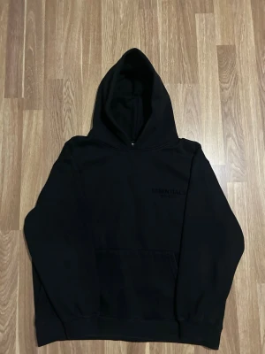 Svart Essentials hoodie med huva - Svart hoodie från Essentials med stor huva och känguruficka framtill. Diskret Essentials-logga på bröstet och större logga på ryggen. Oversized passform och mjukt material som känns riktigt skönt. Perfekt för en avslappnad streetwear-look.