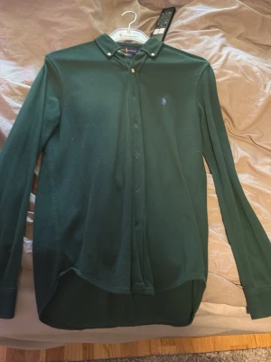 Mörkgrön skjorta med knappar - Snygg mörkgrön skjorta med klassisk krage och knappar hela vägen framtill. Skjortan har lång ärm och en liten broderad logga på bröstet. Perfekt till jeans eller chinos för en stilren look.