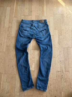 Nudie jeans - Säljer ett par klassiska blå slim fit jeans med fem fickor och snygga slitningar framtill. Jeansen har normal midja och raka ben. Materialet är denim i bomull och passformen är smal. Perfekta för en avslappnad och trendig look.