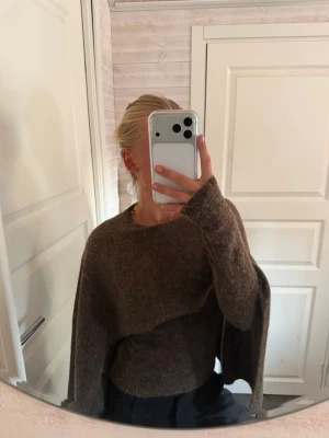 Brun oversized stickad tröja - Mysig brun stickad tröja med oversized passform och långa ärmar. Tröjan har en unik asymmetrisk design med extra tyg över ena axeln, vilket ger en cool och avslappnad look. Perfekt för lager på lager och höstmys.