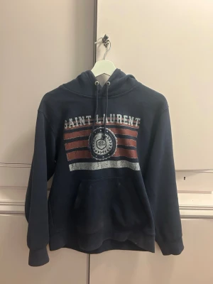 Blå Saint Laurent hoodie med tryck - Mörkblå hoodie från Saint Laurent med huva och dragsko. Framsidan har ett vintageinspirerat tryck med texten 'Saint Laurent' och röda samt vita ränder. Mjuk insida och känguruficka framtill. Perfekt för en avslappnad streetwear-look. Tvättar in den skickas!