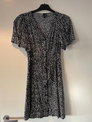 Svartvit prickig klänning Vero Moda - Kort svart klänning från Vero Moda med vita prickar, v-ringad hals och knytband i midjan. Klänningen har korta, lösa ärmar och ett flowy fall som ger en avslappnad men snygg look. Perfekt för sommardagar eller när du vill känna dig extra fin.