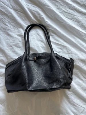 Svart handväska i skinnimitation - Snygg svart handväska i skinnimitation med två rejäla handtag och strukturerad yta. Väskan har en mjuk, rymlig form och minimalistisk design utan synliga loggor. Perfekt för dig som gillar stilrena accessoarer.