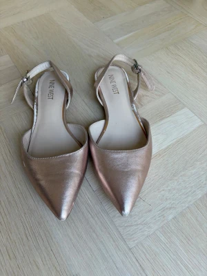Roséguldiga sandaler från Nine West - Säljer ett par stilrena sandaler från Nine West i roséguldigt material. Skorna har spetsig tå, öppen häl och justerbart spänne runt ankeln. Perfekta för dig som vill ha en elegant touch till din outfit. Det har en liten slitning (se sista bild) :(