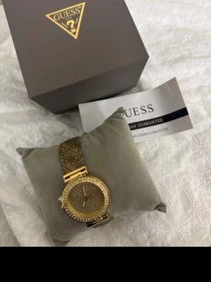 Guldig klocka från Guess - Snygg guldig klocka från Guess med glittrande stenar runt urtavlan och ett mönstrat armband i guld och brunt. Klockan har en rund form och klassisk design, perfekt för dig som vill sticka ut med en elegant accessoar.