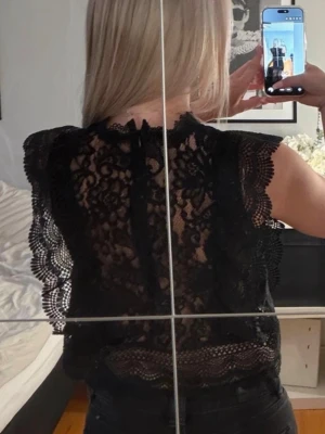 Svart spetsblus med volangdetaljer - Säljer en svart blus i spets med vackra volangdetaljer längs ärmar och nederkant. Blusen är ärmlös och har ett genombrutet mönster som ger en elegant look. Perfekt att styla med både jeans och kjol.