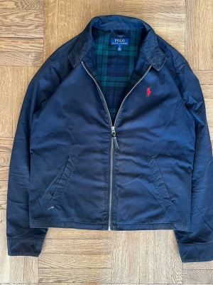 Marinblå Harringtonjacka från Polo - Klassisk marinblå Harringtonjacka från Polo Ralph Lauren med röd broderad logga på bröstet och rutigt foder i blått och grönt. Jackan har dragkedja framtill, snedställda fickor och en stilren krage. Perfekt för en avslappnad men snygg look.