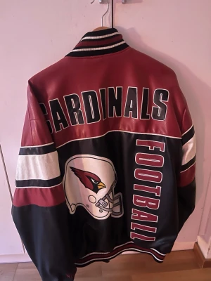 Cardinals bomberjacka i skinnimitation - Vintage NFl Säljer en snygg bomberjacka med Cardinals-tryck och stor football-logga på ryggen. Jackan är vinröd och svart med vita detaljer, har ribbade muddar och krage samt breda ränder på ärmarna. Materialet är syntet/skinnimitation och jackan har en loose passform.