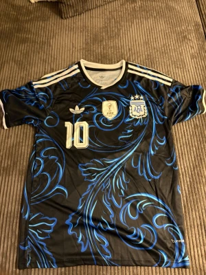 Argentina Messi tröja svart/blå - Snygg svart Argentina fotbollströja med blått mönster, nummer 10 och Messi på ryggen. Tröjan har vita detaljer, AFA-logga, FIFA-märke och klassiska ränder på axlarna. Perfekt för dig som älskar fotboll och vill sticka ut på planen eller läktaren.