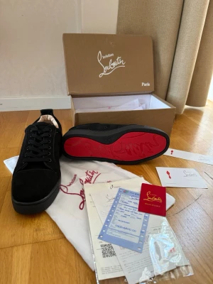 Svarta Louboutin sneakers med röd sula - Snygga svarta sneakers från Christian Louboutin med ikonisk röd sula och diskret logga. Skorna har en klassisk låg modell med svarta skosnören och ovandel i mocka. Perfekta för dig som vill sticka ut med exklusiv stil och detaljer.
