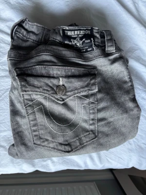 Gråa True Religion jeans bootcut - Säljer ett par gråa jeans från True Religion med lila detaljer. Sitter jätte bra men har sytt in i midjan.