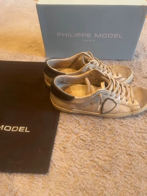 Philippe Model beige sneakers - Snygga vita sneakers från Philippe Model med vita snören. Skorna har en klassisk låg siluett och är tillverkade i läder med diskret logga på sidan. Perfekta för en trendig och avslappnad stil. Mycket bra kvalité på skorna, köpte dom för ca 5000 och ett snöre har lossnat lite på toppen men kan lätt fixas, hälarna har även blivit väldigt nötta men dom är fixade hos en skomakare så nu är dom som vanligt bara inte i samma färg.