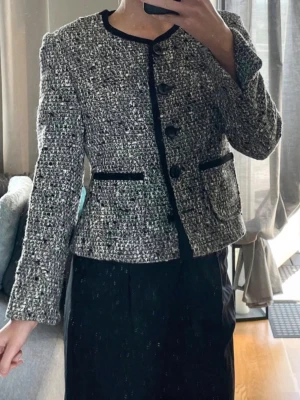 Grå tweedkofta med svarta detaljer - Trendig gråmelerad tweedkofta med svarta kanter och stora knappar framtill. Koftan har två fickor med svarta detaljer och en rak, kortare passform. Materialet är grovt vävt och ger en exklusiv känsla. Perfekt för dig som gillar klassisk men modern stil.