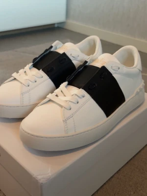 Valentino open svart - Snygga vita sneakers från Valentino Garavani med bred svart läderdetalj över snörningen. Klassisk låg modell med vita skosnören och diskret logga på plösen. Tillverkade i slätt läder för en clean och modern look.