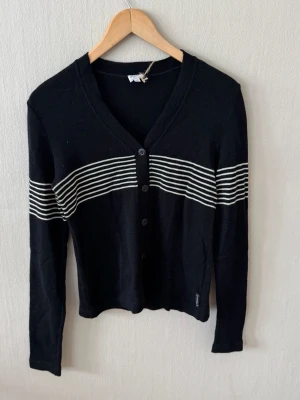 Armani Jeans Svart kofta med vita ränder - Snygg svart kofta med smala vita horisontella ränder över bröstet och armarna. Koftan har v-ringning, knäppning framtill med fyra knappar och långa ärmar. Perfekt till lager på lager och enkel att matcha med olika outfits.0076