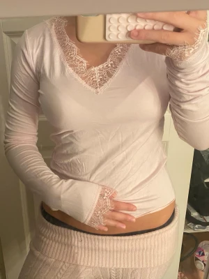 Ljusrosa långärmad topp med spets - Söt ljusrosa långärmad topp med v-ringning och fina spetsdetaljer vid halsringning och ärmslut. Toppen är tight i modellen och har en mjuk, stretchig känsla. Perfekt för dig som gillar romantiska och feminina plagg.