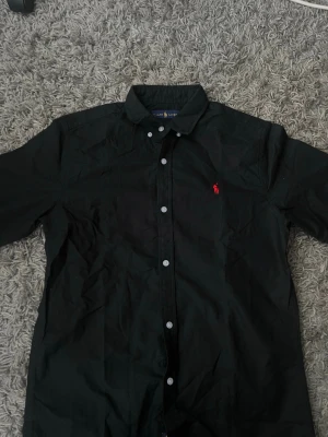 Svart Ralph lauren skjorta med röd logga - Säljer en svart Ralph lauren skjorta med klassisk krage och knappar framtill. Skjortan har en liten röd broderad logga på bröstet och är tillverkad i ett mjukt bomullstyg. Perfekt för dig som gillar stilrena plagg med en enkel detalj. Den är en L men passar som en M 