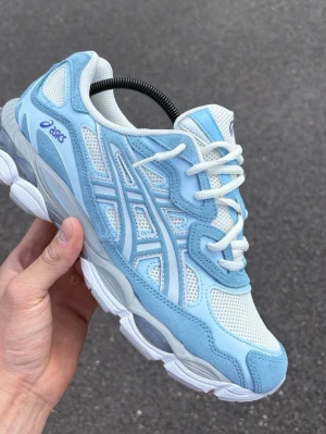Nya oandvända Asics blåvita sneakers mesh & mocka - Snygga sneakers från Asics i ljusblått och vitt. Ovandelen är i mesh och mocka för en sportig vibe, med vita snören och detaljer i lila. Sulan är chunky och ger bra stöd. Perfekta för dig som gillar streetwear och retrostil.
