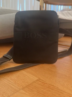 Svart axelväska från Hugo Boss sällsynt!! - Snygg svart axelväska från Hugo Boss med präglad logga på framsidan. Väskan har en fyrkantig form och justerbar axelrem. Tillverkad i ett slätt material som ger en stilren look. Perfekt för att bära det viktigaste med stil.