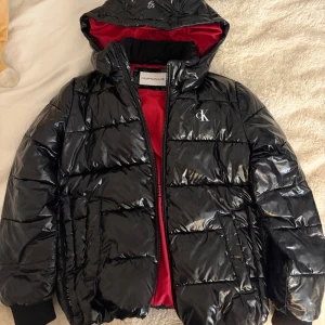 Svart glansig puffer jacka med röd insida - En svart glansig puffer jacka med röd insida från Calvin Klein Jeans. Den passar mig som normalt bär XS och S. 
