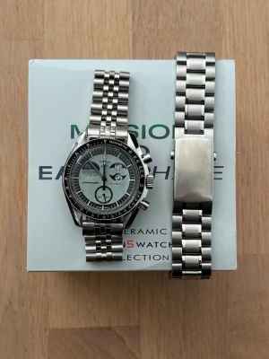 Moddad Omega x Swatch Mission to Earthphase - Detta är en omega x swatch Moonswatch mission to earthphase som är modifierad med en metallboett och metallarmband inspirerat av omega speedmaster. Perfekt för dig som vill ha en moonswatch men tycker att det känns fjantigt med en plastklocka eller drömmer om en speedmaster. Den har en skön tyngd och är byggd på korrekt sätt med delar av hög kvalitet. 42mm stor. Låda, original boett och extra länkar följer med. Skriv om du har några frågor! 