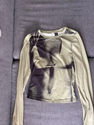 Beige långärmad topp från SHEIN - Trendig beige långärmad topp från SHEIN med ett coolt grafiskt tryck framtill i svart. Tunn och stretchig med figurnära passform, perfekt att styla med jeans eller kjol. Materialet känns mjukt och följsamt mot huden.