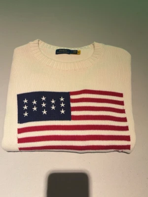 Vit stickad tröja med USA-flagga - Vit stickad tröja från Polo Ralph Lauren med en stor amerikansk flagga i rött, vitt och blått på bröstet. Tröjan har rund halsringning, ribbade muddar och RL-broderi nedtill. Perfekt för dig som gillar klassisk och preppy stil.