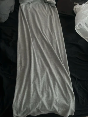 Gray Maxi Dress on Bed - This image shows a long, gray, sleeveless maxi dress laid out flat on a bed. The fabric appears soft and slightly shiny, possibly made of a jersey or modal blend. The dress is gathered at the top and bottom, giving it a relaxed, draped look.