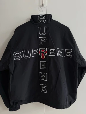 Supreme Jacket - Snygg svart vindjacka från Supreme med stort vitt SUPREME-tryck på ryggen och ett rött R-märke. Framsidan har ett broderat klubbmärke och Supreme-logga på bröstet samt vit piping. Jackan har hög krage och resår vid ärmslut och midja.