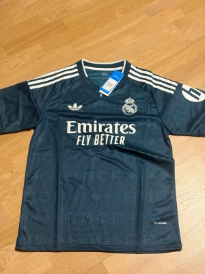 Real madrid 26/27 borta M - Snygg mörkblå Real Madrid matchtröja från Adidas med vita detaljer och klubbmärke på bröstet. Tröjan har korta ärmar, rund hals och tre vita ränder på axlarna. Tillverkad i lätt och ventilerande material med Climalite-teknologi.