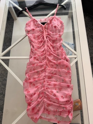 Rosa blommig ruched klänning - Supersöt rosa klänning med tunna axelband och ruched design framtill. Klänningen har ett blommigt mönster i rött och vitt samt en justerbar snörning vid bysten. Materialet ser ut att vara lätt och luftigt, perfekt för varma dagar.