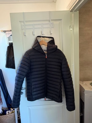 dunjacka Save The Duck M - Snygg svart dunjacka från Save The Duck i storlek M. Jackan har huva, dragkedja framtill och är quiltad med horisontella sömmar. Tillverkad av 100% djurfria material och har ljus insida. Perfekt för kalla vinterdagar.