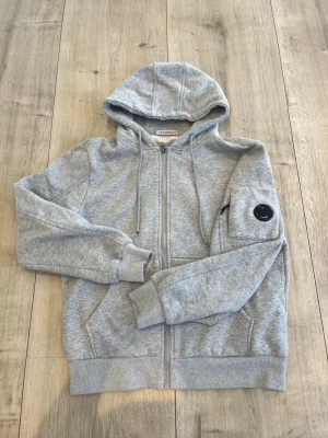 Grå hoodie från C.P. Company - Snygg grå zip-hoodie från C.P. Company med huva och dragsko. Klassisk design med fickor framtill och ikonisk svart linsdetalj på ärmen. Tillverkad i mjuk bomullsblandning som känns skön mot huden. Perfekt för en avslappnad streetwear-look.