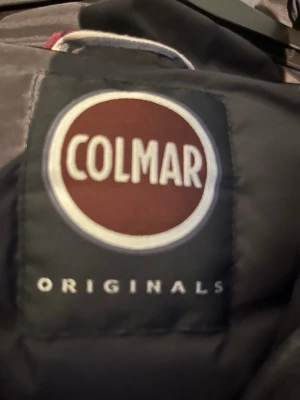 Svart dunväst från Colmar - Snygg svart dunväst från Colmar med quiltat mönster och hög krage. Västen har två dragkedjeförsedda fickor framtill och en logga på bröstet. Perfekt att slänga över en hoodie för en chill look. Materialet är lätt och värmande.