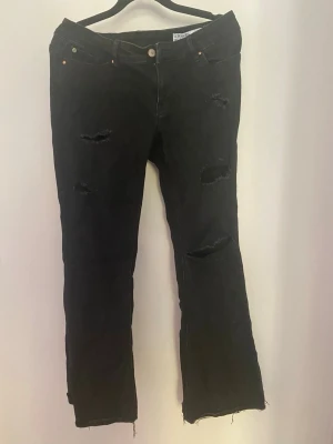 Svarta slitna jeans med bootcut - Säljer ett par svarta jeans med bootcut och slitningar på både lår och knän. Jeansen har råa, fransiga benslut och klassisk femficksmodell. Perfekta för en edgy och avslappnad stil.