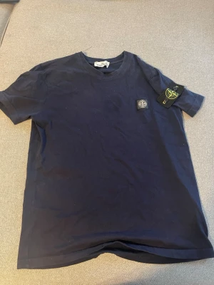 Mörkblå t-shirt från Stone Island - Snygg mörkblå t-shirt från Stone Island med klassisk logopatch på bröstet och en avtagbar logga på ärmen. T-shirten har rund hals och är tillverkad i mjuk bomull för skön komfort. Perfekt för dig som gillar streetwear och stilrena plagg.