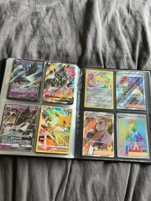 Pokémonkort samling i pärm - Säljer en pärm med Pokémonkort, inklusive flera holografiska och full art-kort som Necrozma, Pikachu, Blastoise och Professor's Research. Kort i olika färger och motiv, många med glansig finish och unika illustrationer. Perfekt för samlare eller Pokémonfans. Får med bindern
