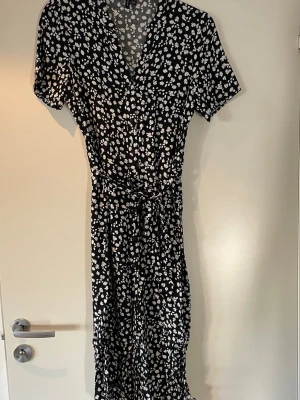 Svart blommig klänning Vero Moda - Svart långklänning från Vero Moda med vitt blommigt mönster. Klänningen har v-ringning, korta ärmar och ett knytband i midjan som ger en snygg siluett. Perfekt för dig som gillar en feminin och avslappnad stil.