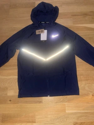 Mörkblå Nike vindjacka med reflex - Snygg mörkblå vindjacka från Nike med huva och dragkedja. Jackan har en tydlig reflexdetalj i V-form över bröstet och en vit Nike-logga. Perfekt för löprundan eller när du vill synas i mörkret. Lätt och smidig design.