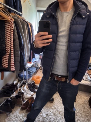 Mörkblå pufferjacka med huva Cain - Säljer en mörkblå pufferjacka med huva och dragkedja framtill. Jackan har ribbade ärmar och sidofickor med dragkedja. Perfekt för kalla dagar och har en modern, sportig look som passar till jeans och sneakers.