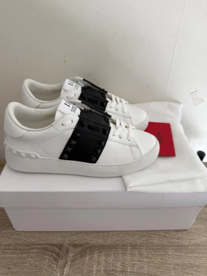 Vita sneakers med svarta nitar - Snygga vita sneakers med en bred svart rem över vristen dekorerad med svarta nitar. Skorna har en chunky sula och en modern, stilren design. Perfekta för dig som vill sticka ut med detaljer och gillar streetwear-inspirerad stil.