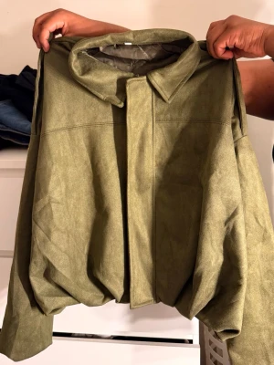 Olivgrön bomberjacka med krage - Snygg olivgrön bomberjacka med klassisk krage och dold dragkedja framtill. Jackan har en oversized passform och är fodrad på insidan för extra komfort. Materialet ser ut som mockaimitation och ger en cool, modern look.