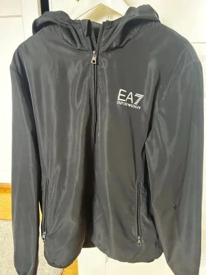 Svart vindjacka från EA7 Emporio Armani - Snygg svart vindjacka från EA7 Emporio Armani med huva och dragkedja framtill. Jackan har två sidofickor med dragkedja och logga på bröstet. Perfekt för blåsiga dagar och enkel att matcha till olika outfits. OBS: litet brännhål på ärmen! Kan lätt fixas  med lagningslapp Pris kan DISKUTERAS!