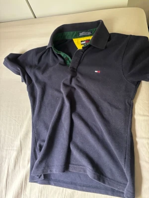 Mörkblå pikétröja Tommy Hilfiger - Klassisk mörkblå pikétröja från Tommy Hilfiger med korta ärmar och liten logga på bröstet. Kragen har gröna och gula detaljer på insidan för en cool kontrast. Perfekt till jeans eller shorts för en avslappnad look.